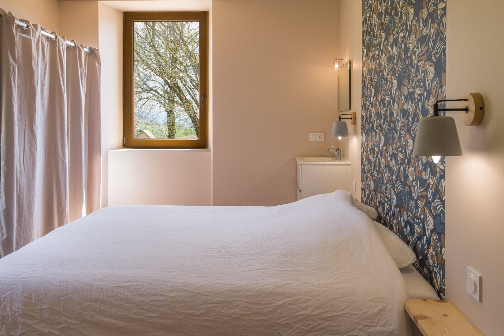 Een slaapkamer met een bed, een raam, een nachtkastje, een lamp en een muur met een bloemenprint.