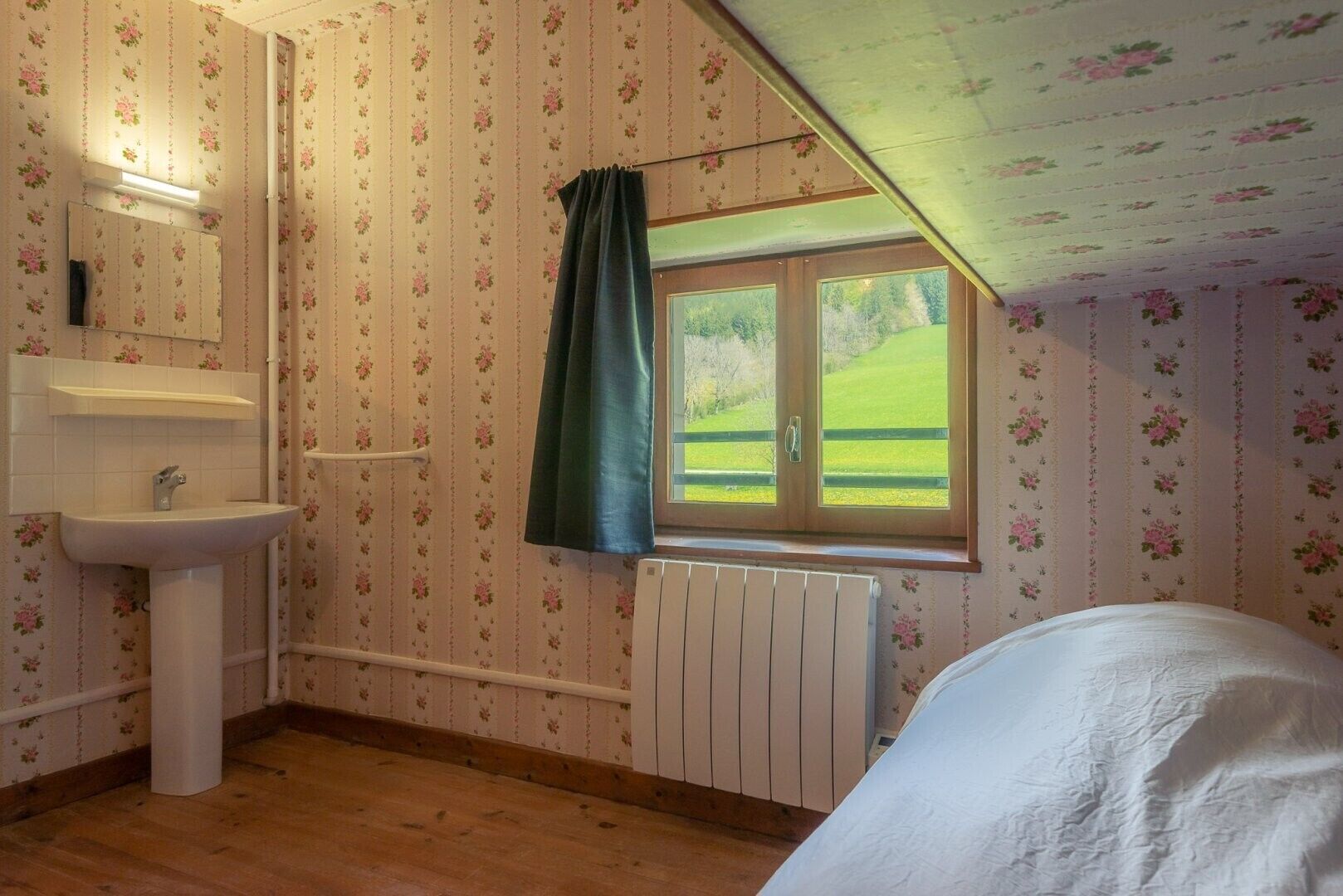 Un dormitorio con piso de madera, una ventana con vistas a un paisaje verde, una cama blanca y un baño con lavamanos y espejo.