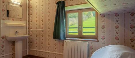 Un dormitorio con piso de madera, una ventana con vistas a un paisaje verde, una cama blanca y un baño con lavamanos y espejo.