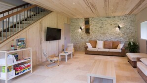 Een woonkamer met een houten trap, een stenen muur, witte meubels en een aan de muur bevestigde televisie.