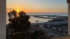 Une vue sur le coucher de soleil depuis un balcon donnant sur une marina, avec des bateaux et une digue incurvée.