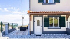Une maison avec une terrasse carrelée, une porte blanche et des volets verts.