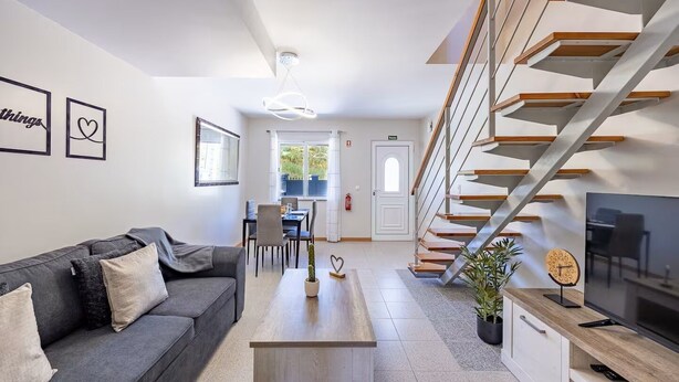 Un salon moderne avec un canapé gris, une table basse en bois et un téléviseur posé sur un meuble en bois. Un escalier avec des marches en bois et des rampes en métal mène à une autre pièce.