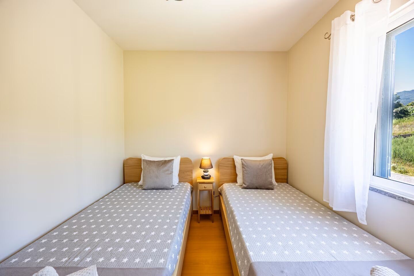 A small, single-room accommodation with two single beds, a bedside table with a lamp, and a window with a view of greenery.