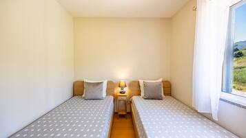 A small, single-room accommodation with two single beds, a bedside table with a lamp, and a window with a view of greenery.