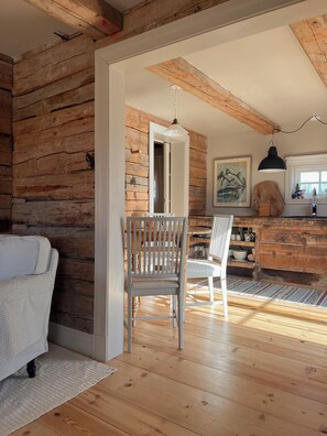 A rustic interior with exposed wooden walls, a dining area with a wooden table and chairs, a kitchen with a hanging light, and a framed artwork on the wall. - Snickarboden – Cozy farmhouse living room near the woods and the lake (Kronobergs län)