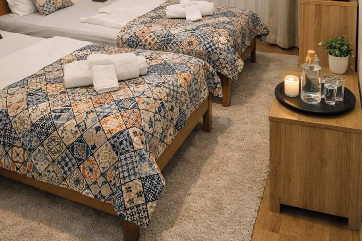 A hotel room with two beds, a bedside table with a lamp, a TV mounted on the wall, and a small table with a candle and glasses.