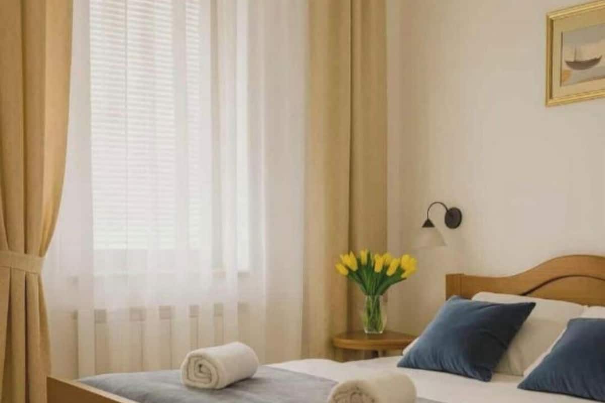 A neatly made bed with a blue blanket and two pillows, a vase with yellow flowers on a bedside table, and a painting on the wall.