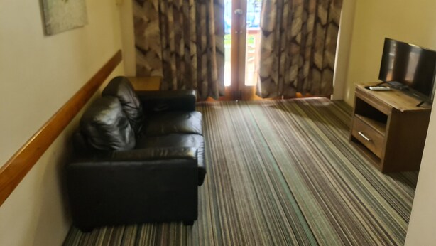 A room with a black leather sofa, a wooden TV stand with a flat-screen TV, and a patterned carpet. - Cairns holiday unit apartment kitchen aircon wifi near esplanade and hospital (Cairns North)