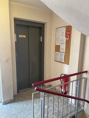 A hallway with a closed elevator door, a bulletin board with notices, a fire extinguisher, and a metal railing. - Little Oasis Harz - Wi-Fi - Balcony (Braunlage)