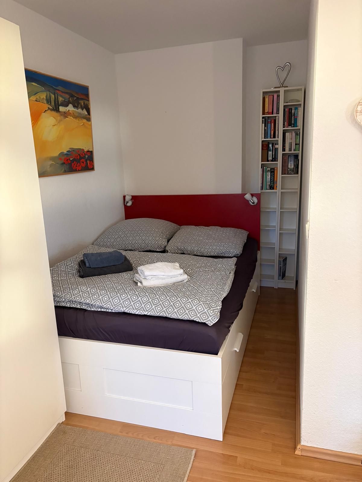 A bedroom with a bed, a bookshelf, and a painting on the wall.