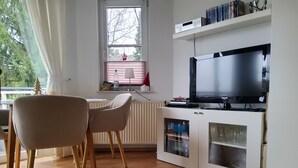 Un salon moderne avec un téléviseur à écran plat, des chaises en bois, un radiateur et une fenêtre donnant sur des arbres.
