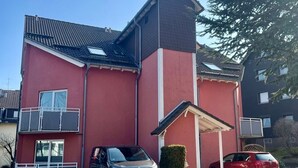 Un immeuble de deux étages avec une façade rouge, un toit en tuiles et un petit balcon. Deux voitures sont garées devant, et un arbuste est visible sur la droite.