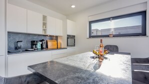 Een moderne keuken met witte kasten, een zwart aanrechtblad en een ingebouwde oven. Er is een zithoek met een tafel en stoelen, en een raam met uitzicht op een gebouw.