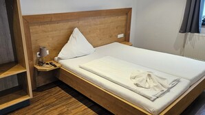Un llit ben arrenjat amb capçal de fusta, una taula de nit amb una làmpada i un comandament a distància, i un moble prestatge integrat.