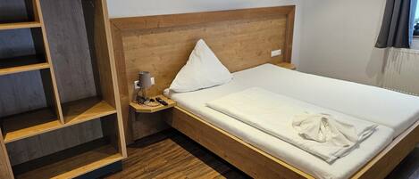 A neatly made bed with a wooden headboard, a bedside table with a lamp and a remote control, and a built-in shelf unit.