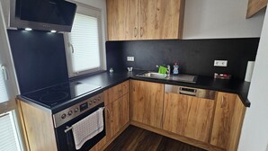 Una cuina compacta amb fogó negre, armaris de fusta, forn integrat, un rentapeus i una finestra amb persianes.