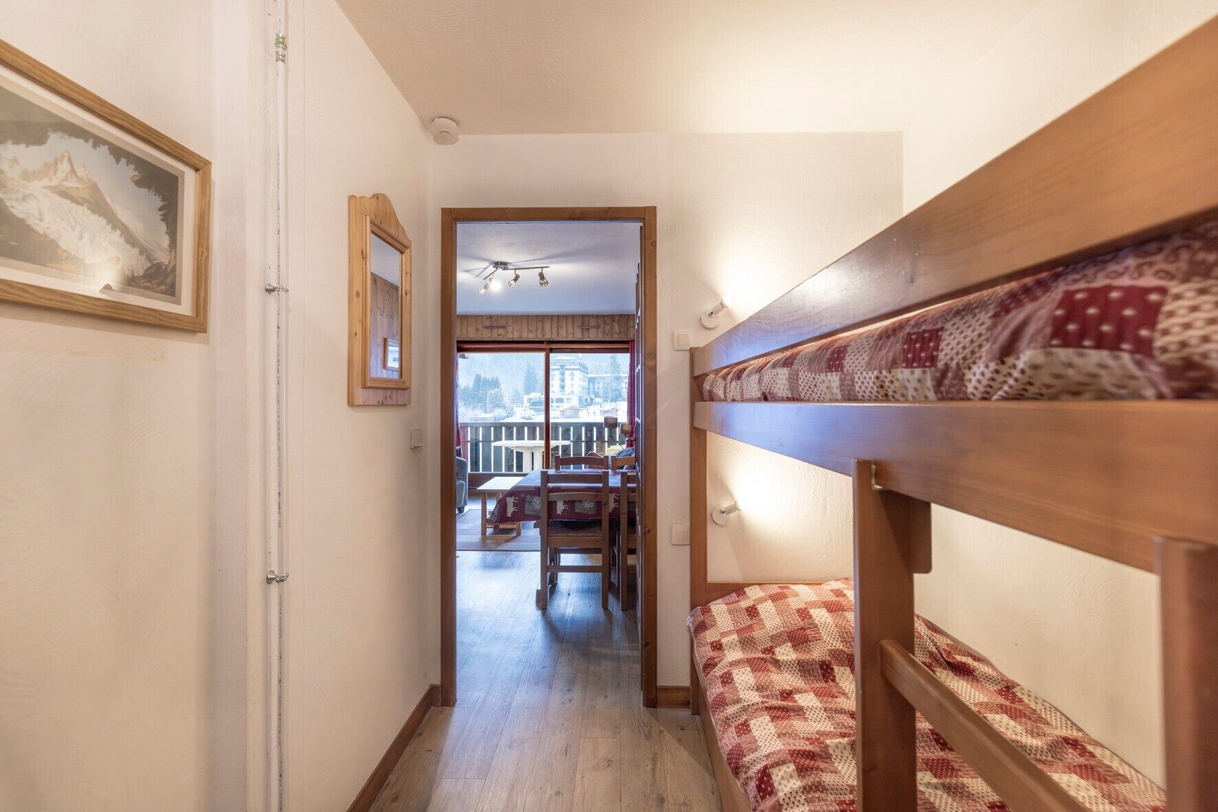 Un corridoio stretto con un letto a castello di legno sulla destra, una porta a sinistra e vista su un'area pranzo con tavolo e sedie.