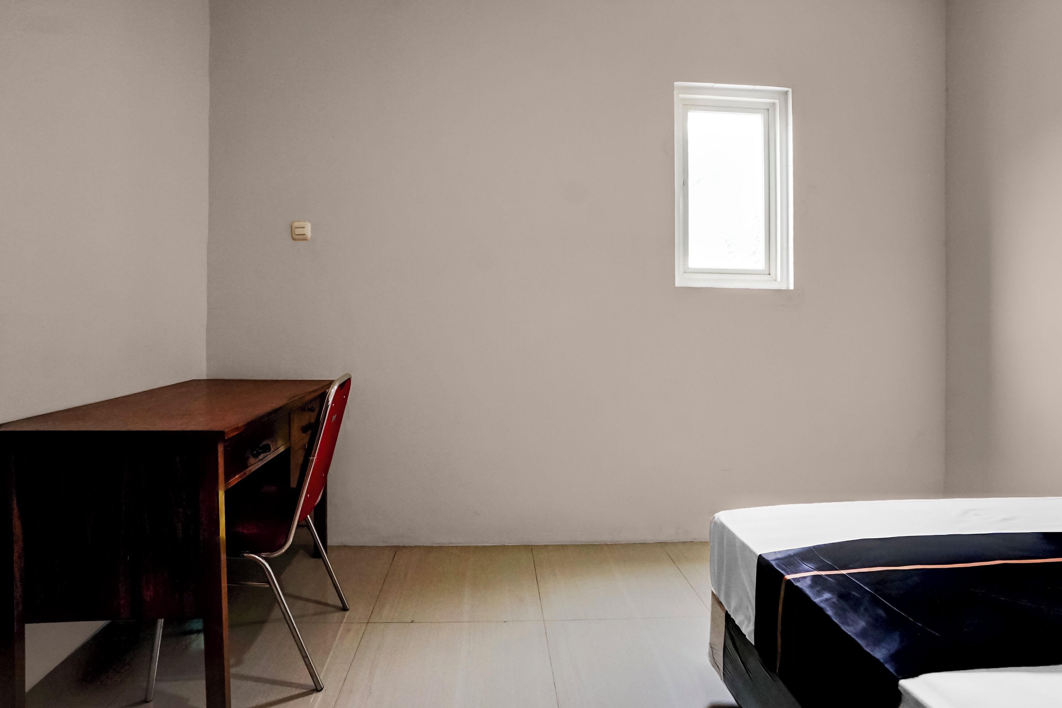 A room with a wooden desk, a red chair, a bed with a white mattress, and a small window.