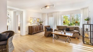 A living room with a sofa, a coffee table, and a wicker chair. There is a cabinet with decorative items and a window with curtains. - Apartment Gartenblick, 96 sqm, 2 Bedrooms, Max. 4 Guests (Balve)