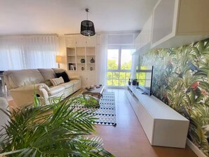 Ein Wohnzimmer mit einer Couch, einem Couchtisch, einem Fernseher und einem Bücherregal. Pflanzen und ein Fenster mit Blick auf Bäume sind ebenfalls vorhanden.