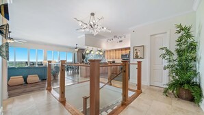 A spacious living area with a glass railing, a chandelier, and a large window overlooking a balcony. - Sailmakers Place 903 | Two-Story Penthouse | Panoramic Views | My Beach Getaways (Pensacola)
