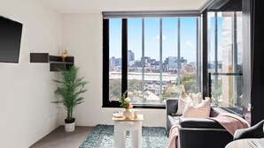 A modern living room with a large window offering a city view, a potted plant, a small table with a vase, and a sofa with cushions. - Comfy City Apt Beside Queen Victoria Market (Melbourne)