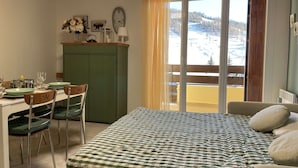 Ein Zimmer mit einem Bett, einem Esstisch mit Stühlen, einem grünen Schrank und einem Fenster mit Blick auf eine verschneite Landschaft.