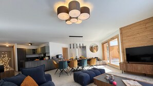 Una sala de estar moderna con un sofá, un reposapiés, una mesa de centro y una televisión. Hay un comedor con sillas y una mesa, y una cocina al fondo.
