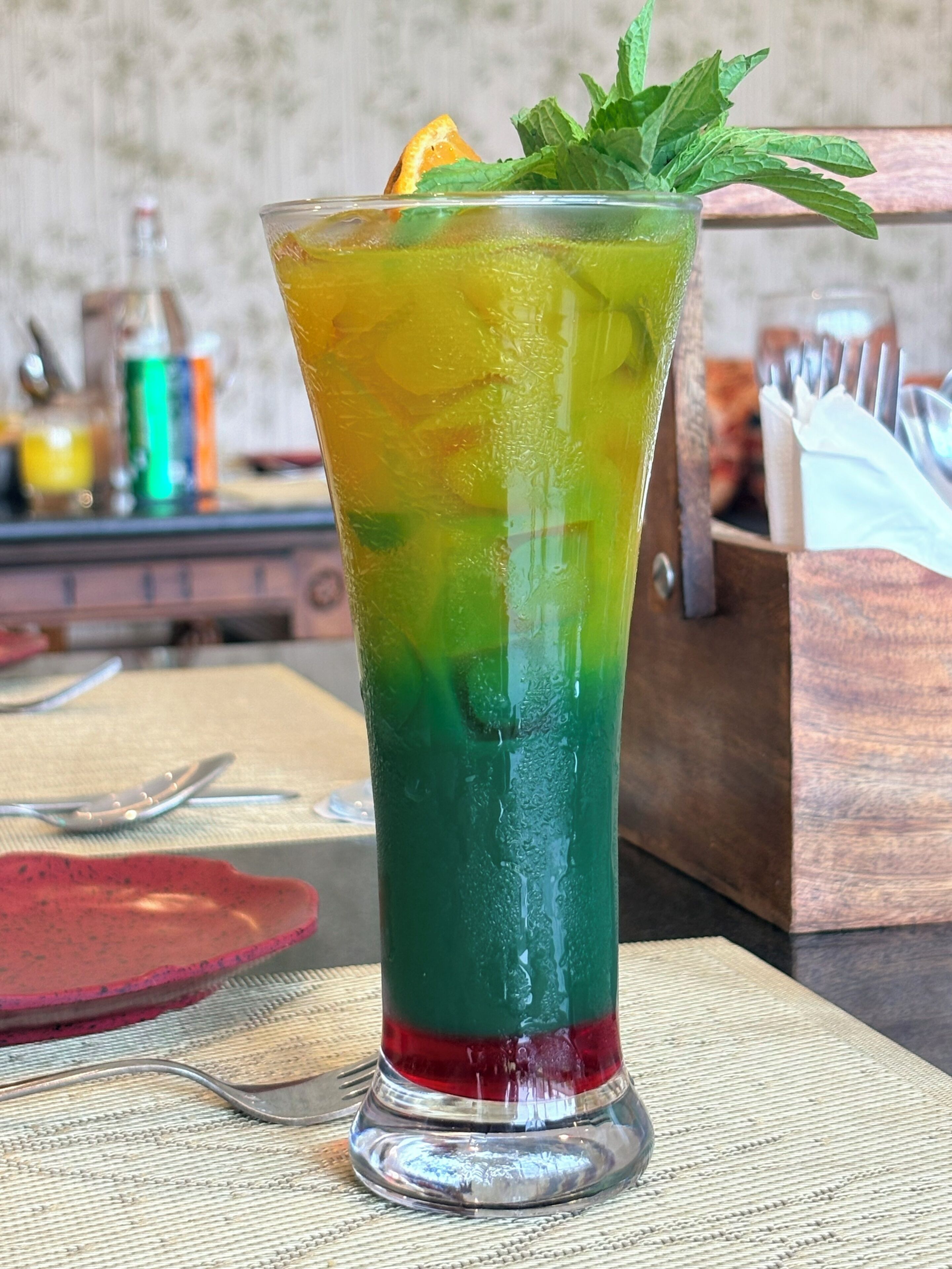 A tall glass with a layered drink, garnished with mint and an orange slice, on a table with a placemat.
