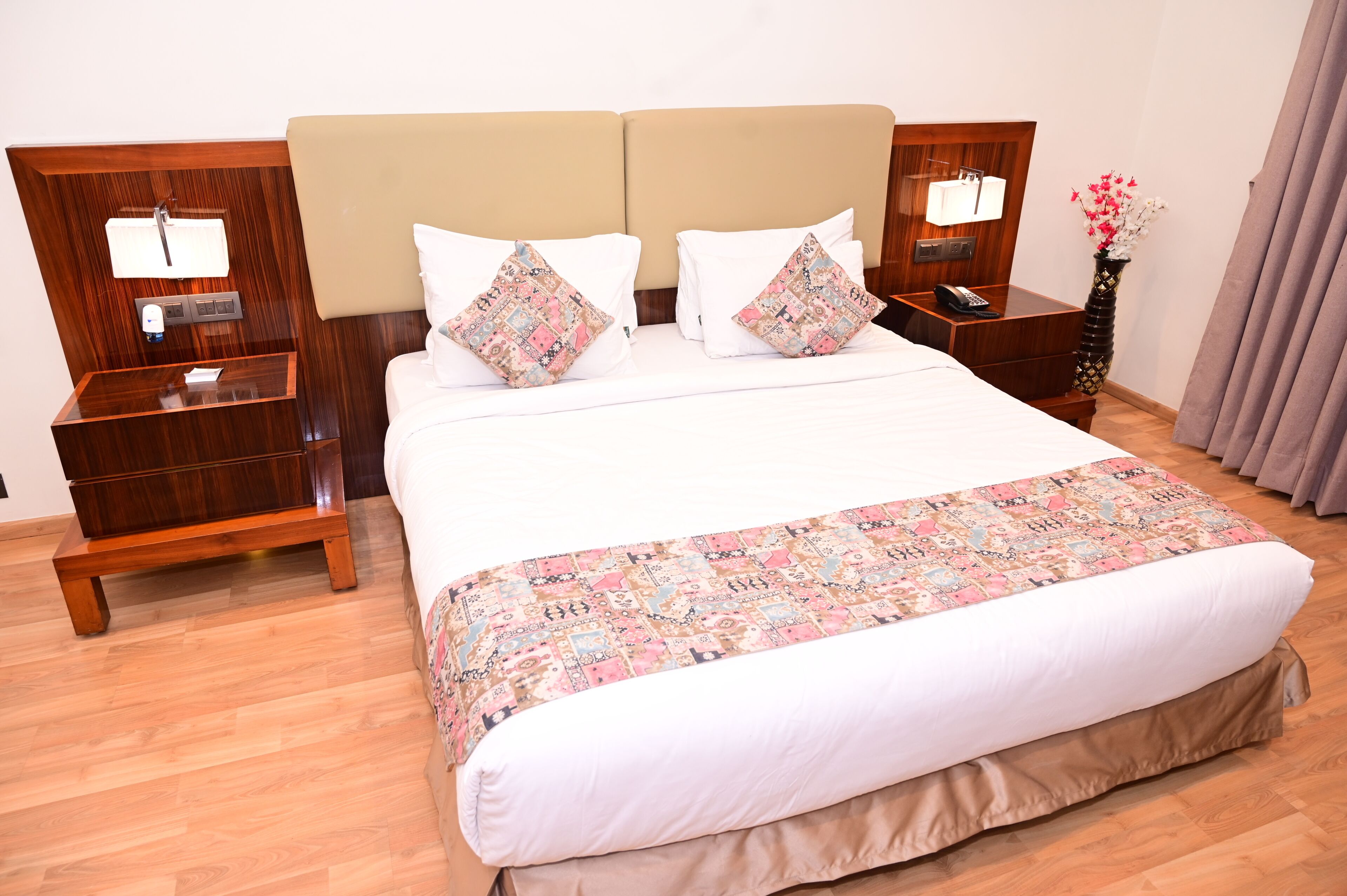 A hotel room with a large bed, two bedside tables, a nightstand, a bedside lamp, a vase with flowers, and a window with curtains.