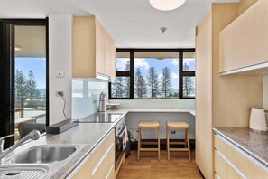 A modern kitchen with a large window, wooden stools, and a view of trees. - Bright Coastal 3BR Apartment Opposite the Beach (Sunshine Coast)