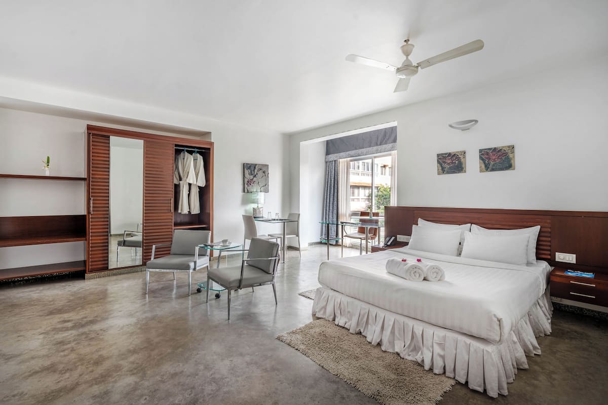 A spacious bedroom with a large bed, a desk, and a chair. There is a wardrobe, a ceiling fan, and artwork on the walls.