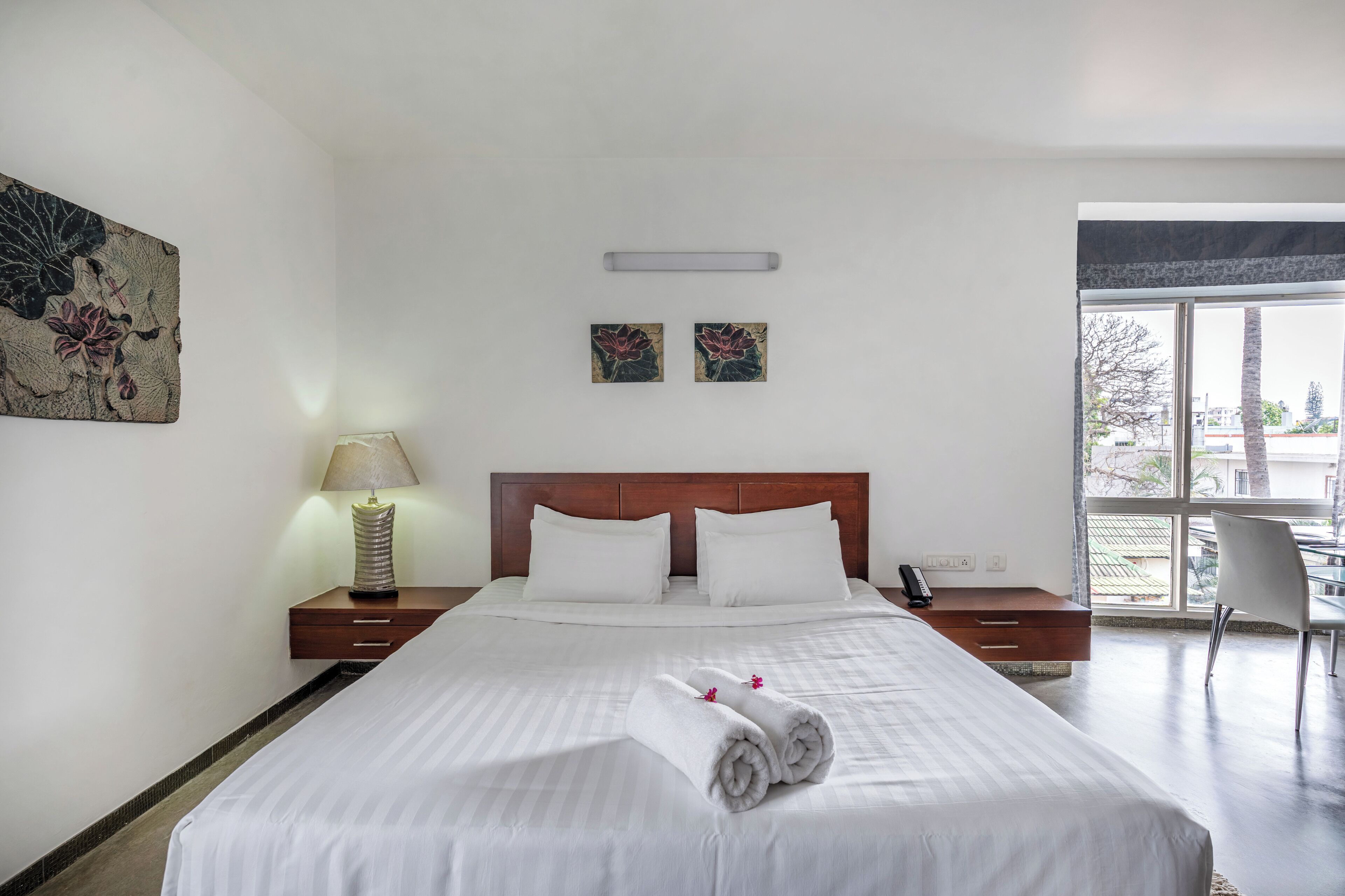 A hotel room with a large bed, a nightstand with a lamp, two wall-mounted paintings, and a view of the outside through a large window.