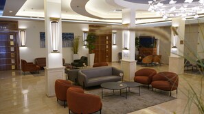 A modern lobby with a grey sofa, orange armchairs, and a round coffee table. There are potted plants and a wooden cabinet in the background. - Aristocrat Hotel (Bene Beraq)