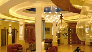 A hotel lobby with a modern design, featuring a central seating area with sofas and a chandelier. There are wooden doors and a large mirror on the wall. - Aristocrat Hotel (Bene Beraq)