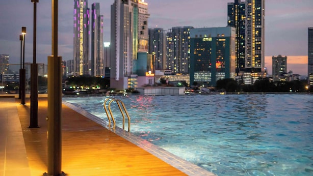 A waterfront pool with a wooden deck and metal railings, surrounded by high-rise buildings with illuminated windows. - (Kids Fun) 2BR Space Apt near KSL CIQ JB (Johor Bahru)