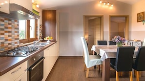 Eine moderne Küche mit Gasherd, Spüle und Holzschränken. Ein Essbereich mit Tisch und Stühlen bietet einen Blick ins Freie durch ein Fenster.