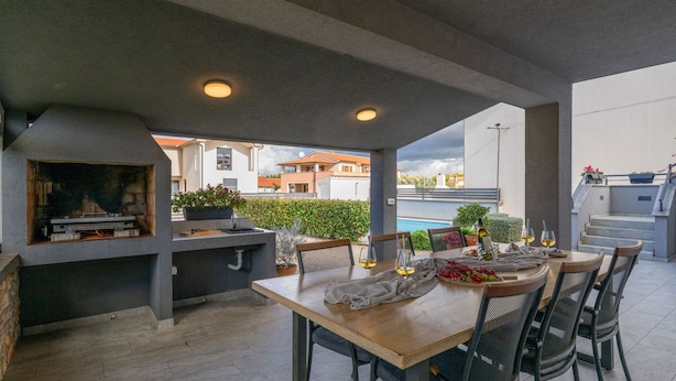 Een terras buiten met een barbecue, eettafel en stoelen, met uitzicht op een zwembad en woongebouwen.