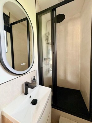 A modern bathroom with a white sink, a round mirror, and a glass-enclosed shower. - La Villa Bligny  (Étretat)