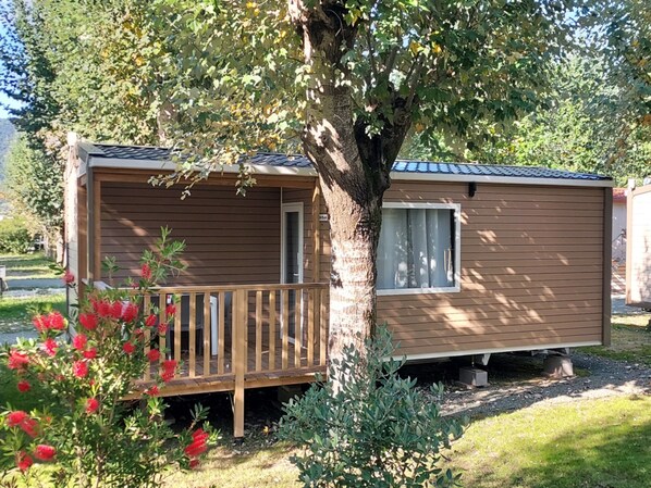 A small, single-story cabin with a wooden deck and a window, surrounded by greenery and a tree. - Mobile Home in Sarazena in Einem Ferienpark (Sarzana)