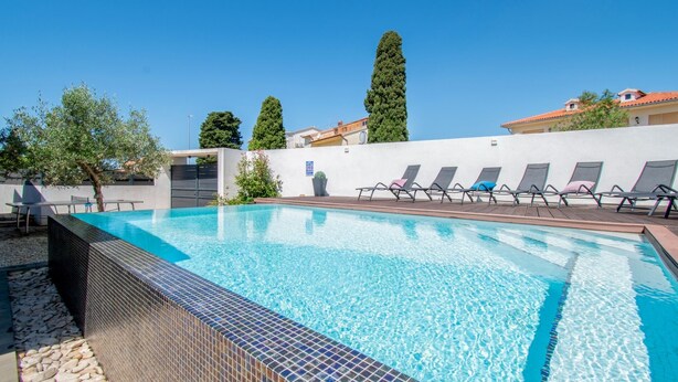 Ein Schwimmbecken mit gefliesten Rändern, Liegestühlen und einer weißen Wand, im Hintergrund Bäume und ein Gebäude.