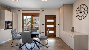 A modern kitchen with an integrated oven, a dining area with a round table and four chairs, a large wall clock, and a view through a glass door to an outdoor patio. - Kuca za odmor Hana by Villas Guide (Vodnjan)