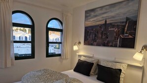Et hotelværelse med en seng, to vinduer med buet top, et bybillede-maleri og et natbord med lamper.