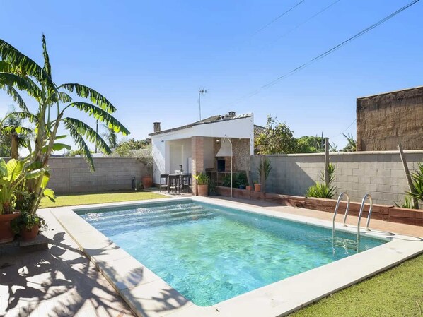 A rectangular swimming pool with a white deck, surrounded by a low brick wall and a grassy area. There is a covered patio area with a table and chairs, and a small brick building in the background. - Villa Near Puntalejo Beach (Conil de la Frontera)