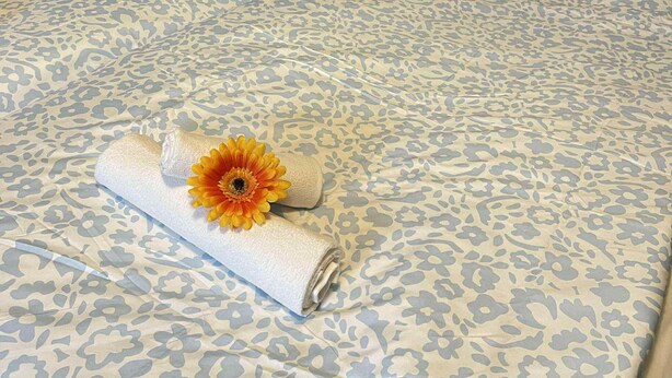 A neatly made bed with floral patterned bedding, a rolled white towel, and a yellow flower. - Nymans pensionat (Aneby)