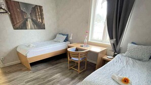 A compact hotel room with two single beds, a small desk with a chair, and a window with curtains.