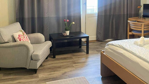 A bedroom with a bed, a sofa, a small table with a vase of flowers, and a desk with a computer.