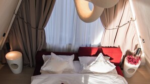 A bed with white and red bedding, a small bedside table with a bottle and flowers, and a window with curtains.