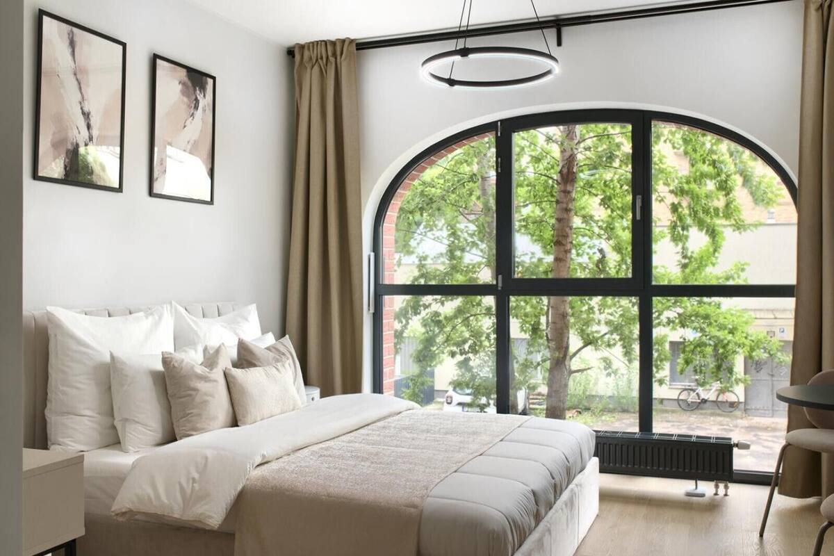 Une chambre moderne avec une grande fenêtre en arc de cercle, un lit agrémenté de nombreux coussins et deux tableaux abstraits accrochés au mur.